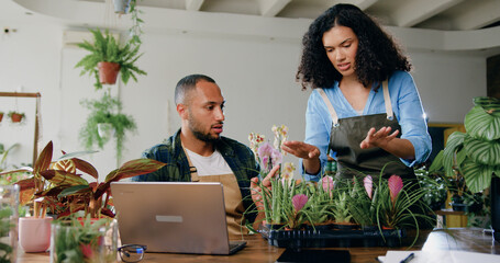 African American smiling man and woman florists in aprons working in flower shop and typing on laptop. Family couple making online service to accepting orders delivery of plant or flowers from a