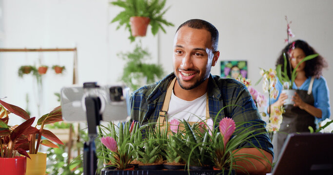 Smiling man social media blogger florist recording floristry video tutorial, shooting vlog in flower store. Male greenhouse worker in apron tells clients about her small business during an online