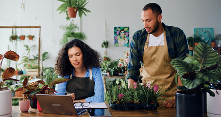 African american woman florist using laptop computer male colleague in apron talking checking...