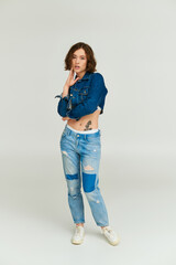 stylish young model with tattoo in cropped denim jacket and blue jeans posing on grey backdrop