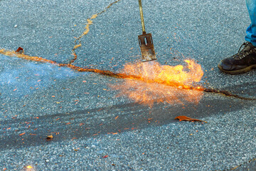 During road surface restoration work worker burns dry grass in asphalt cracks before applying...