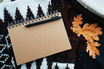 Notepad with black pen on a cozy sweater next to a hat and oak leaves