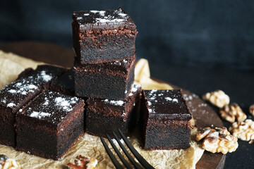 Chocolate brownie next to walnuts and a black fork on a wooden board on a dark background