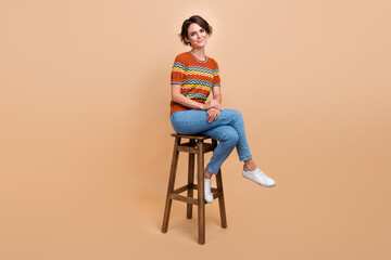 Full size portrait of satisfied glad friendly girl sit wooden stool isolated on beige color...