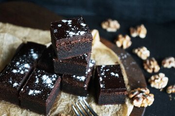 Chocolate brownie next to walnuts and a black fork on a wooden board on a dark background