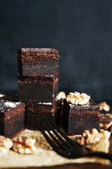 Chocolate brownie next to walnuts and a black fork on a wooden board on a dark background
