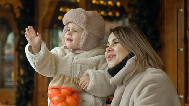 Winter portrait of a young woman with her young daughter at a Christmas fair. The girl and her mother are waving to someone.