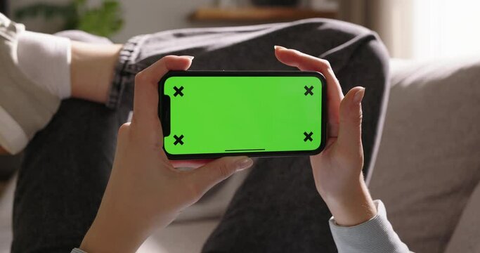 Young woman uses smartphone featuring green screen in landscape mode lounging on sofa. Advertisement of app. Tap on center of screen, favorite movies and videos, modern technologies, entertainment