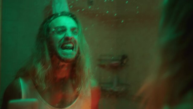 Furious long-haired man screaming emotionally at his reflection in a wet mirror in the bathroom with neon light. Shaky camera shot