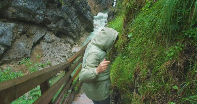 Happy woman walking along the duckboards in rocky gorge. Girl waves her hand and invites you to follow her. Mountain landscape, rainy day.