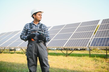 Portrait of Young indian man technician wearing white hard hat standing near solar panels against...