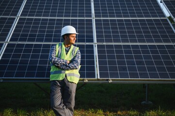 Portrait of Young indian man technician wearing white hard hat standing near solar panels against blue sky. Industrial worker solar system installation