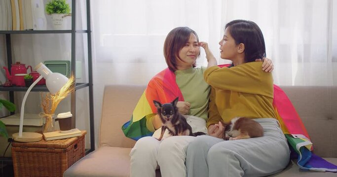 Happy Loving Asian lesbian couple in love smiling looking together while lying on sofa with cute dog at home. couple holding rainbow flag symbol of lgbt, relationship romantic freedom and lifestyle