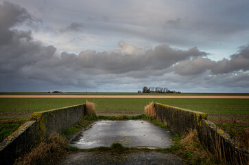 A bridge over the Noordpolder canal in the Noordpolder in the province of Groningen, the Netherlands on the horizon a farm under a cloudy sky.