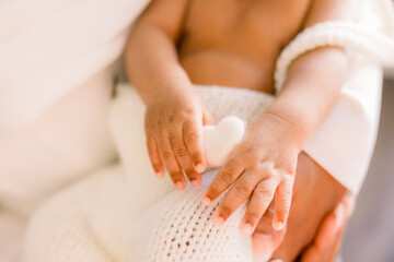 Newborn baby is held in his parents' arms as he holds a heart with his hands. Close-up of his fingers 