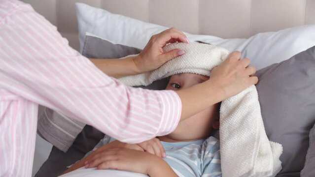 An ill boy feels bad and weak, lies in the bedroom on the bed. A caring young mother sits nearby, makes a compress, applies a wet towel, soaked in cold water, and places it on son forehead to relieve