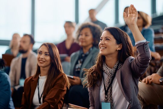 Happy entrepreneur raising her hand to answer question during business conference in convention center.