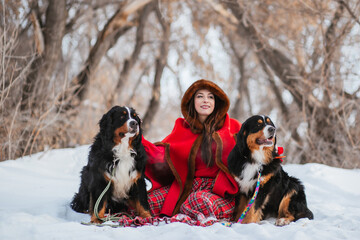 Naklejka na ściany i meble woman in winter in an ancient red coat and plaid skirt in winter in the snow with a dog of the Bernese Mountain Dog breed. Fairytale image of Little Red Riding Hood with a big dog.