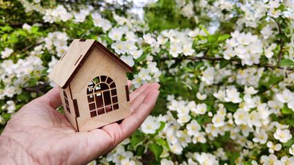 Small wooden toy house on palm of woman hand on natural background with apple flowers. symbol and concept of care, buying, selling, donating of eco friendly home. close-up, soft selective focus