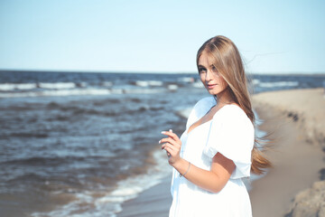 Happy, beautiful woman on the ocean beach standing in a white summer dress