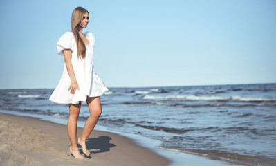 Happy smiling beautiful woman is walking on the ocean beach in a white summer dress