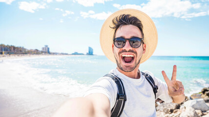 Happy man with hat and sunglasses taking selfie picture with smartphone at the beach - Cheerful traveler having fun outside - Handsome guy smiling at camera - obrazy, fototapety, plakaty