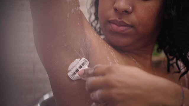 Unrecognizable young black woman shaving her armpits in shower. African American girl removing hair with soft razor standing in bathroom at home. Depilation body care routine and skin cosmetics.