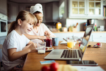 Cute little girl having breakfast with mother using laptop in kitchen
