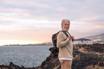 Old senior man walking on the cliff wearing backpack and smiling, enjoying sea excursion at sunset...