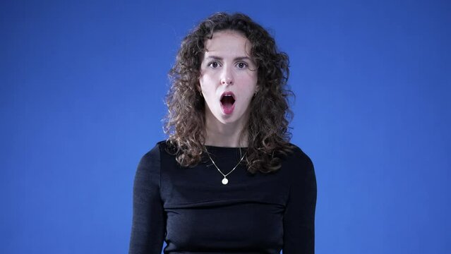 Shocked woman reacting with disbelief in open mouth surprised reaction looking at camera on blue background