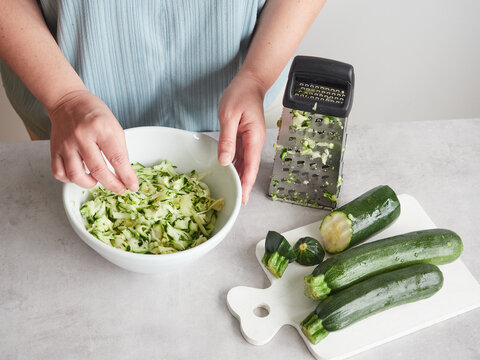 woman with her hands in a bowl of grated zucchini, other fresh zucchini on wooden board and stainless steel hand grater on the countertop