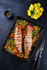 Gebratenes Mangalica Schweinefilet gefüllt mit gehackten Wallnüssen und Kürbis und umwickelt mit Speckmantel serviert mit Gemüse und Salzkartoffel als Draufsicht auf einem Backblech 