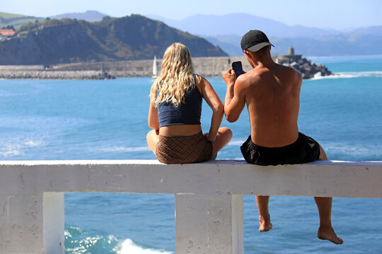 jovenes pareja sentados mirando al mar con m&oacute;vil desde un pretil zumaia verano getaria pa&iacute;s vasco 4M0A2550-as23