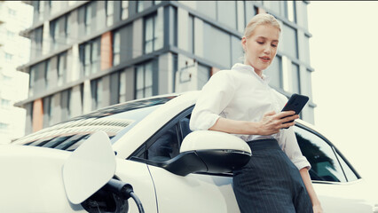 Progressive businesswoman leaning on electric car and charging station before driving around city...