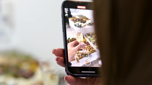 filming food on a phone, close-up, a woman hand with a smartphone films a glass of champagne against the backdrop of buffet snacks, a young unrecognizable woman presses to take a photo