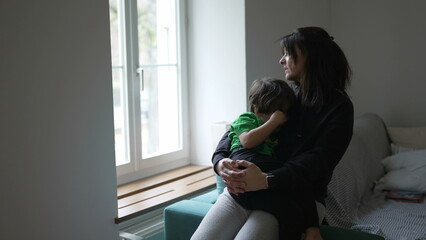 Mother consoling hurt child by window at home. Candid tender moment of thoughtful pensive mom...