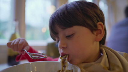 Mother feeding child spaghetti pasta food on bowl covered with napkin on collar. Little boy enjoys...