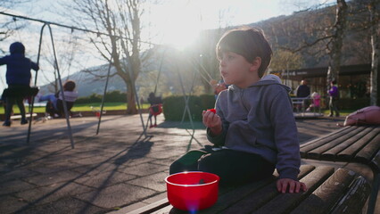 Small boy seated at park snacking while observing his surroundings. Child enjoys food snack at...