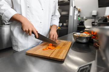 cooking food, profession and people concept - close up of male chef with knife chopping carrot on cutting board at restaurant kitchen