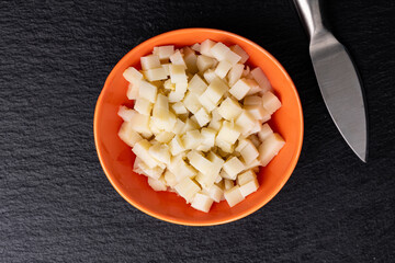 Diced boiled potatoes in orange bowl next to kitchen knife on flat black background.