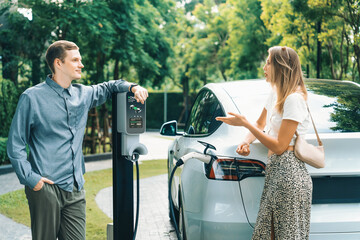 Young couple travel with EV electric car charging in green sustainable city outdoor garden in...