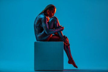 Nude woman with black gloss paint on body sitting on cube on blue background - 676061287