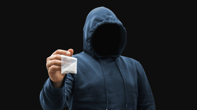 Faceless man in a hood holds transparent plastic bag with white powder hard drugs on dark background, anonymous drug dealer or gangster sells narcotics