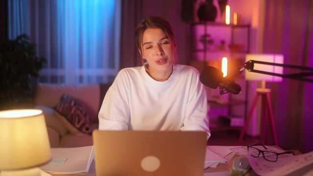 Smiling young woman influencer looking at the camera while recording video blog mobile vlog podcast live stream share experience with audience using microphone sitting at desk at home indoors