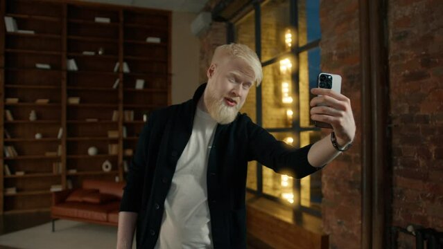 Casual albino businessman taking selfie showing V sign, all good photos on smartphone in loft at night city. Positive man smiling. Handsome hipster showing peace symbol. Guy changing poses for selfie