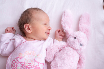 Newborn baby girl smiling while sleeping in bed next to her stuffed rabbit toy.