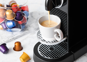 Pouring fresh morning coffee with espresso machine and capsules pods on white marble background.