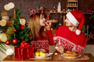 happy lovers a man and a girl in red sweaters in the kitchen with a Christmas tree feed each other...