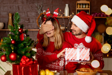 happy lovers a man and a girl in red sweaters in the kitchen with a Christmas tree give each other gifts closing their eyes at home and celebrate New Year or Christmas rejoicing and smiling