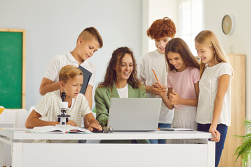 Group of school students and classmates sitting at the desk with microscope in classroom with young woman teacher and looking at the laptop monitor screen during a lesson. Education, science concept.